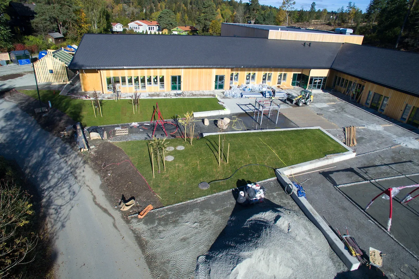 Område under anlegg med ferdiglagte gressplener, små nyplantede trær og lekestativ, plassert ved siden av et nytt bygg med trefasade.